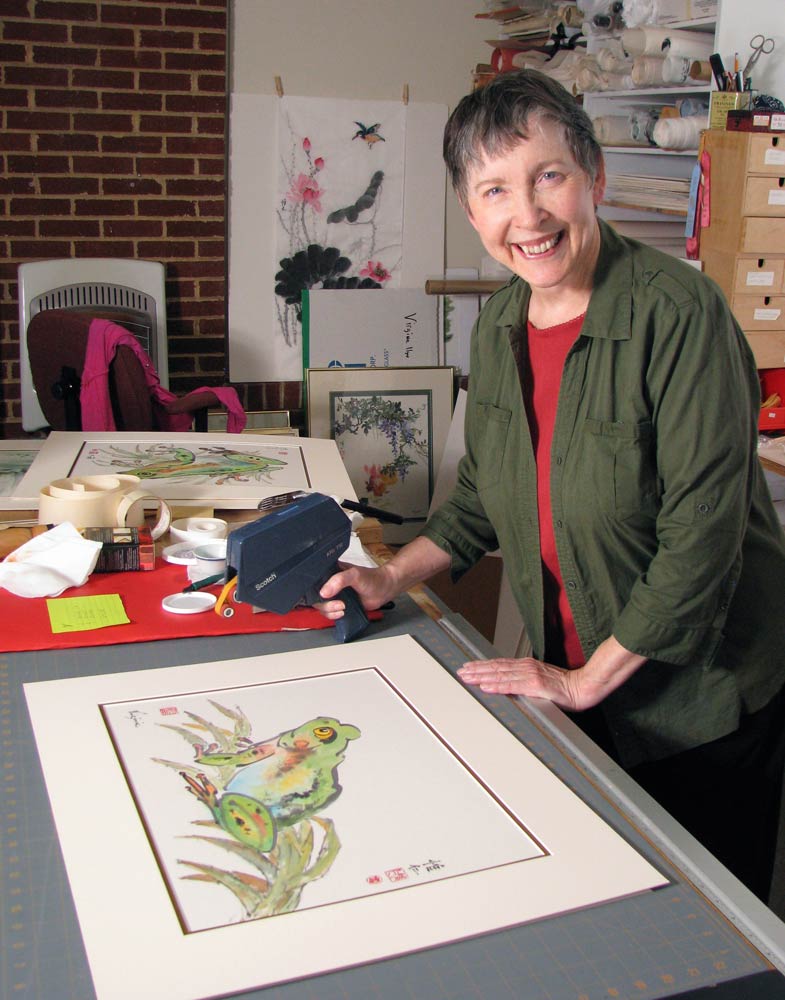 Artist in her studio cutting a mat and applying it to her frog painting. Picture